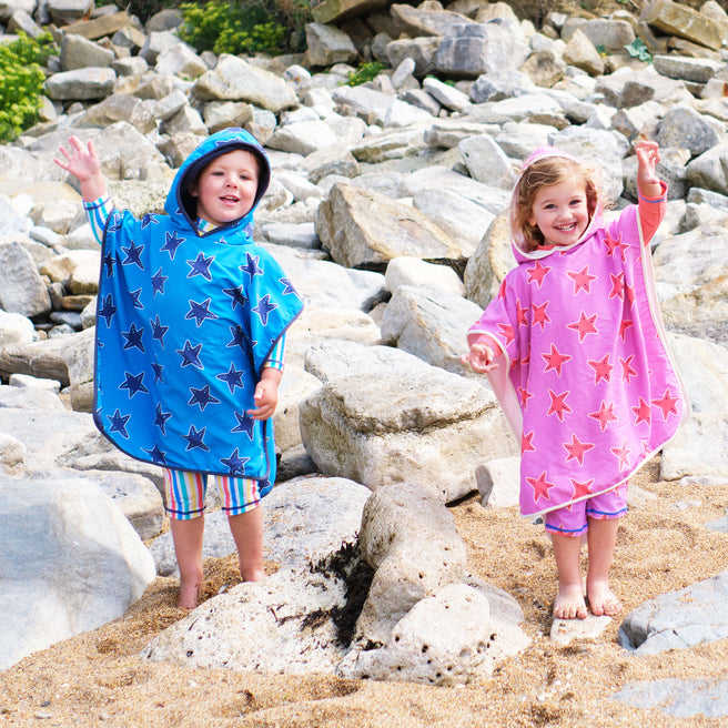 Two children on a beach in hooded Beach Towel Ponchos with star prints, waving to the camera.